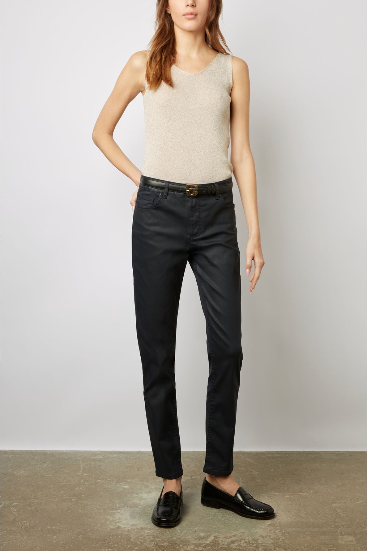 A woman wearing a beige sleeveless top, black belt, dark pants, and black loafers stands against a plain background with one hand in her pocket.