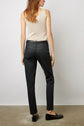 A woman with long hair wears a beige sleeveless top, black pants, and black shoes, standing with her back to the camera against a plain white wall and concrete floor.