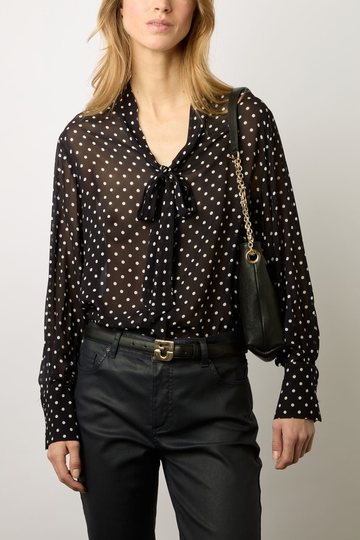 Woman wearing a black sheer blouse with white polka dots and a bow at the collar, black pants, and a black shoulder bag with a gold chain strap, standing against a plain background.