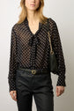 Woman wearing a black sheer blouse with white polka dots and a bow at the collar, black pants, and a black shoulder bag with a gold chain strap, standing against a plain background.