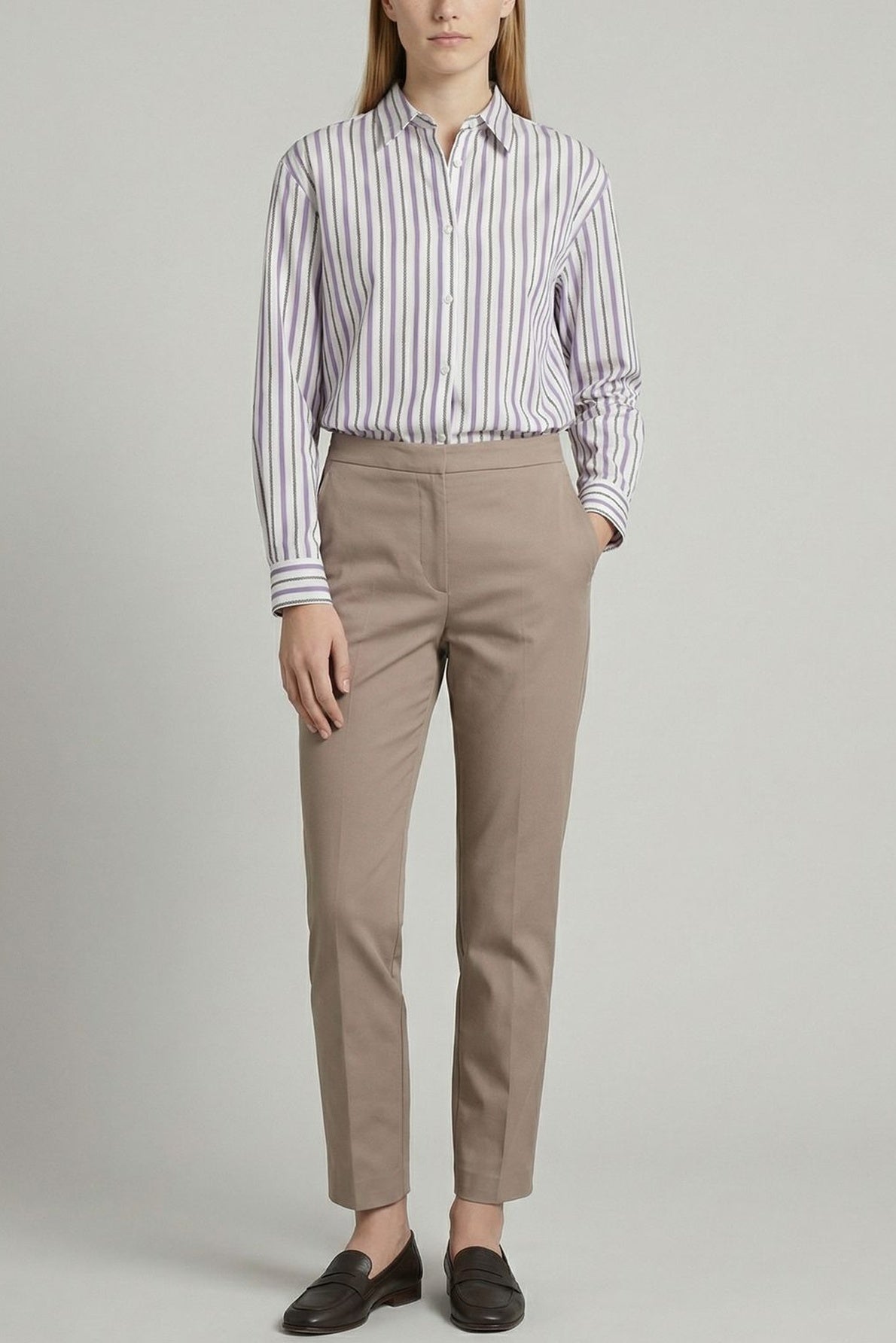 Person standing against a plain background, wearing a striped button-up shirt tucked into beige pants with hands in pockets, and black loafers. Their hair is straight and parted in the middle.