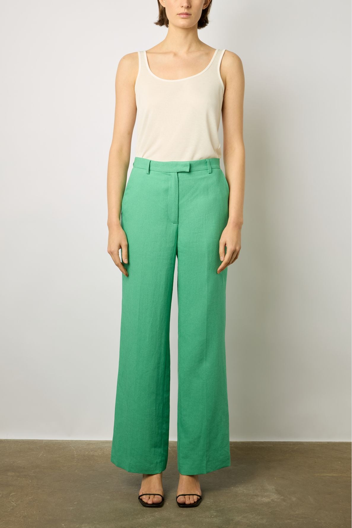 A woman stands against a plain background wearing a white sleeveless top, high-waisted green wide-leg pants, and black open-toe heels.