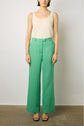 A woman stands against a plain background wearing a white sleeveless top, high-waisted green wide-leg pants, and black open-toe heels.