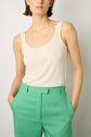 A person wearing a sleeveless white tank top and high-waisted green pants stands with hands in pockets against a plain background.
