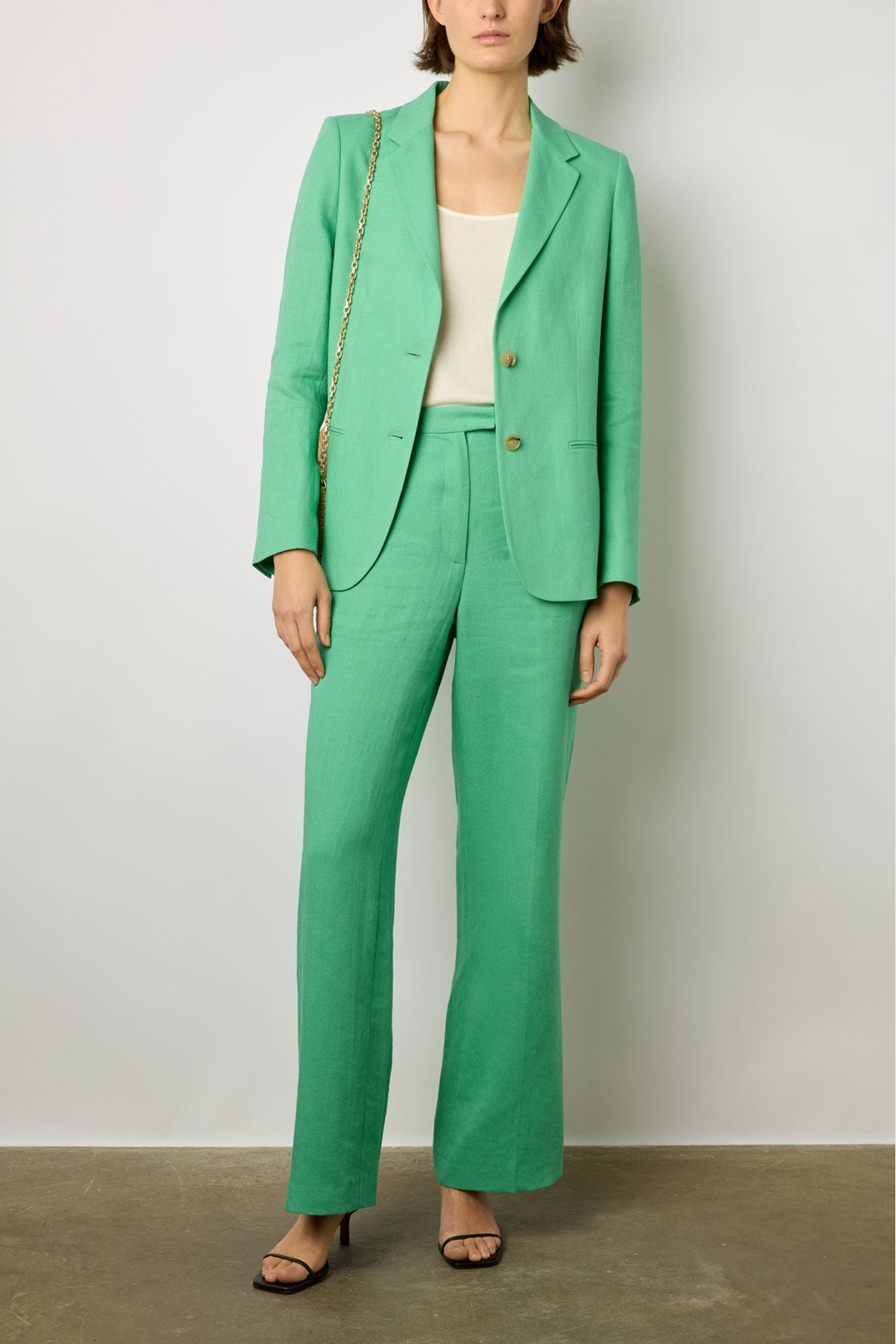 A person wearing a green suit with a white top, black heeled sandals, and a tan chain shoulder bag stands against a plain light gray background. Their face is partially out of frame.