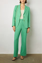 A person wearing a green suit with a white top, black heeled sandals, and a tan chain shoulder bag stands against a plain light gray background. Their face is partially out of frame.