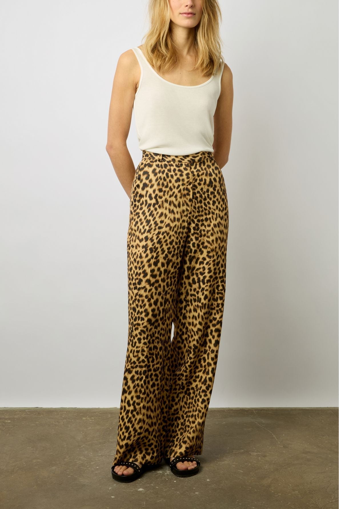 A person wearing a sleeveless white top, high-waisted wide-leg leopard print pants, and black sandals, standing against a plain white background.