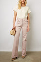 A woman stands against a plain wall wearing a short-sleeve cream top, high-waisted light pink pants, tan heels, and holding a small woven handbag.