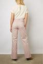 A woman stands with her back to the camera, wearing a short-sleeved cream top and high-waisted, wide-leg light pink pants with brown heeled boots, against a plain background.
