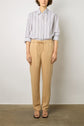 Person standing against a neutral wall, wearing a white button-up shirt with vertical dark stripes, and a coton tencel pantalon large 7/8 with a drawstring waist in tan. Black open-toe sandals complete the look. Hands are relaxed at their sides. Face is not visible.
