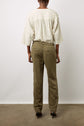 Person standing on a concrete floor facing away, wearing a loose white blouse with thin stripes and olive green pants with an elastic waistband and back pockets, paired with black heeled sandals. Plain white wall in the background.