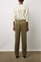 A person stands facing away, wearing an off-white, loose, striped blouse with three-quarter sleeves and high-waisted, olive green pants. The background is plain and the floor is concrete.