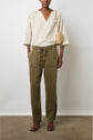 A person wearing a light striped V-neck blouse, olive green drawstring pants, black sandals, and carrying a woven beige shoulder bag stands against a plain background.