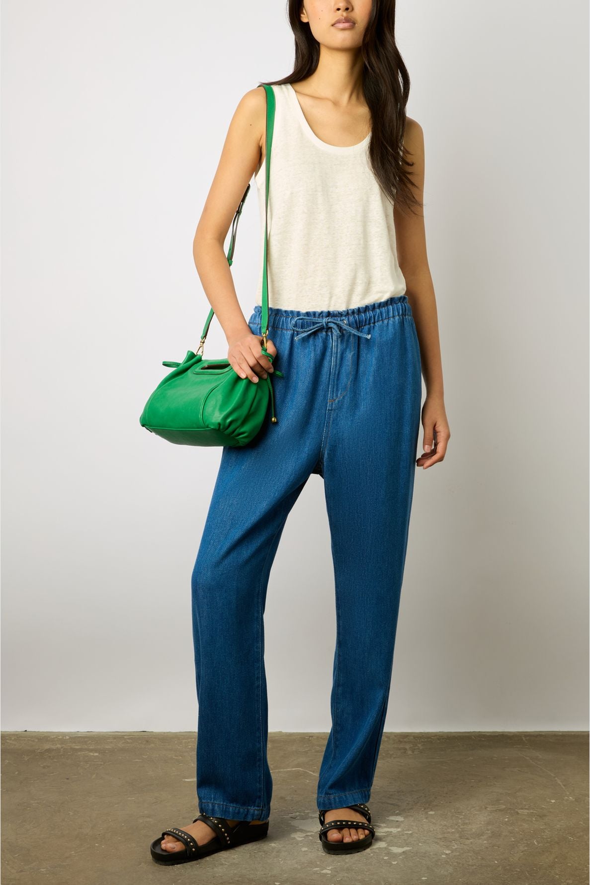 Woman wearing a white tank top, loose blue jeans, black sandals, and carrying a green shoulder bag, standing against a plain background.