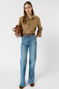 A woman stands holding a brown bag, wearing a tan lace-up shirt, wide-leg blue jeans with visible seams, a detailed belt, and brown shoes, against a plain light background.