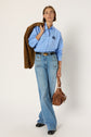 Person in a light blue button-up shirt with a logo, blue high-waisted jeans, black shoes, holding a brown jacket over one shoulder and a brown handbag in the other hand, standing against a plain background.
