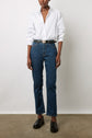 A person stands against a plain wall wearing a white button-up shirt, dark blue high-waisted jeans, a black belt, and black loafers. Their hands are relaxed at their sides.