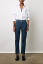 A person stands against a white wall wearing a white button-up shirt, blue jeans, and black loafers. Their hands rest at their sides, and their head is slightly turned, but their face is not fully visible.