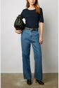 A woman wearing a navy top, high-waisted blue jeans, a black belt, and black clogs stands holding a black handbag. She is posed against a plain, light background.