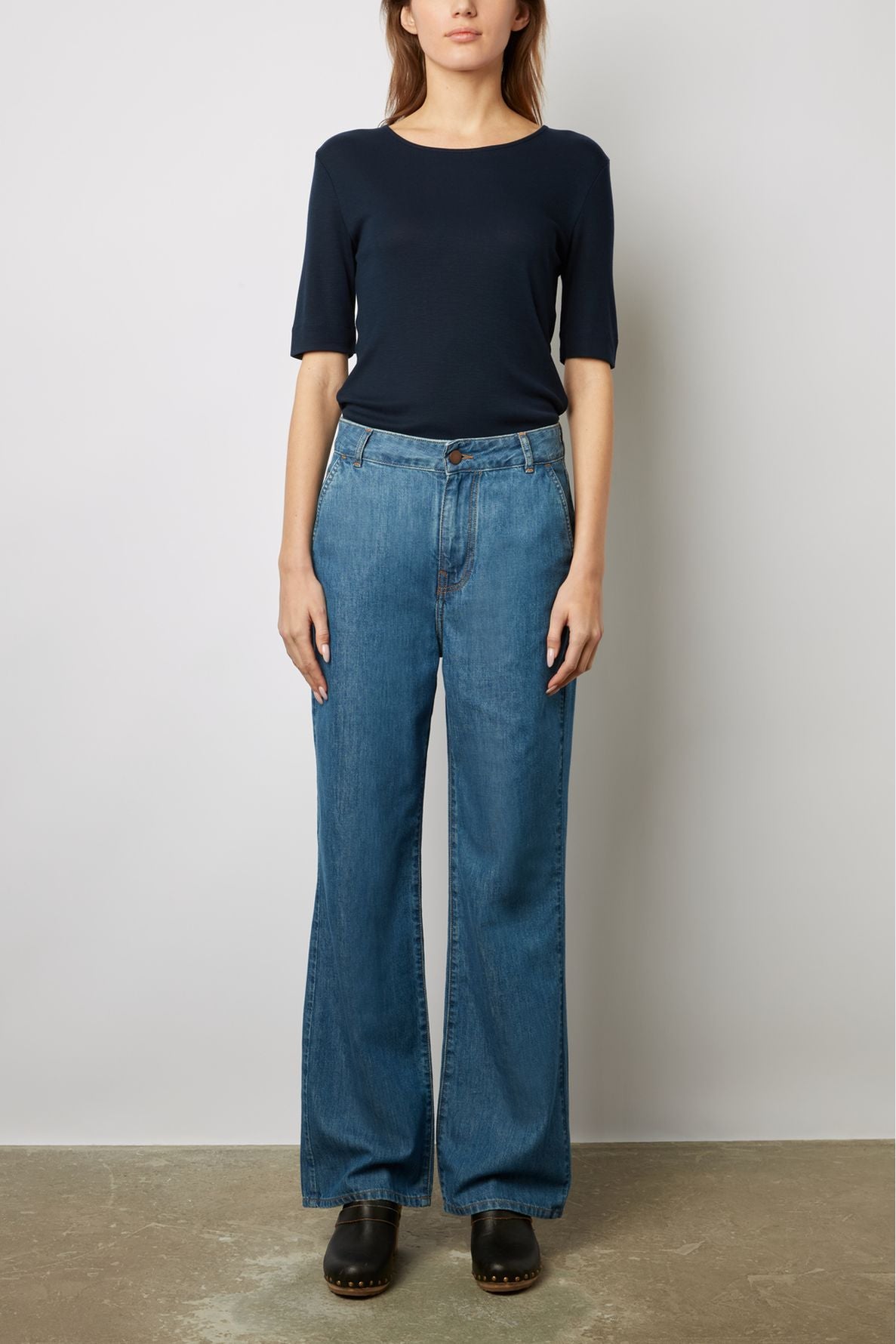 A woman stands against a plain wall, wearing a navy short-sleeve top, high-waisted blue wide-leg jeans, and black clogs.