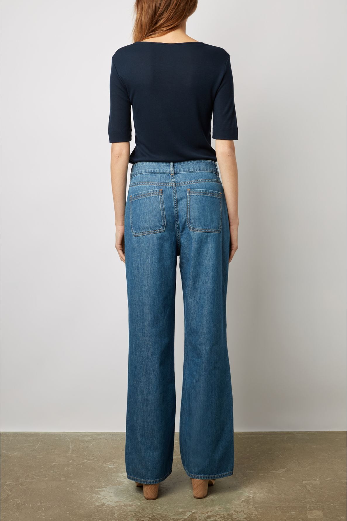 A person with long hair, wearing a navy short-sleeve top and wide-leg blue jeans, stands facing away from the camera against a plain light background.