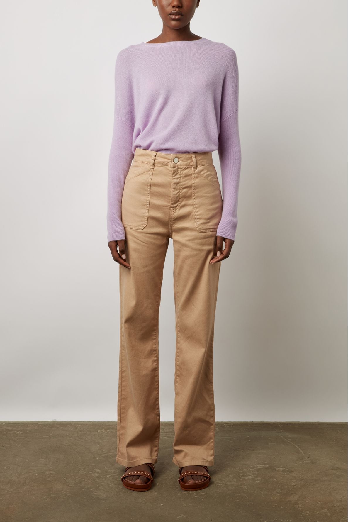 A person stands against a white wall wearing a light purple long-sleeve sweater, high-waisted beige wide-leg pants, and brown sandals. Their hands are relaxed by their sides.