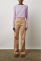 A person stands against a white wall wearing a light purple long-sleeve sweater, high-waisted beige wide-leg pants, and brown sandals. Their hands are relaxed by their sides.