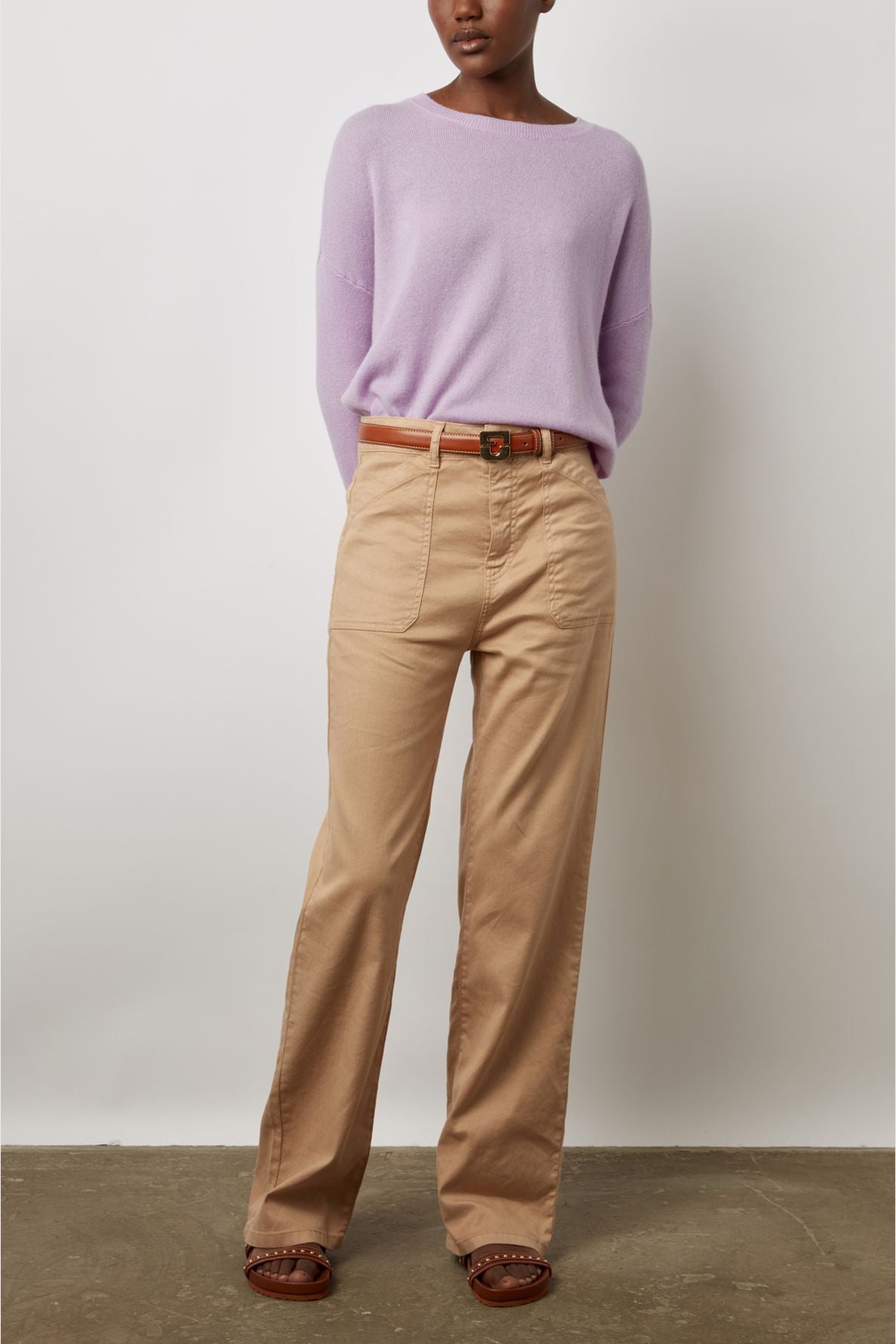 A person stands against a white wall wearing a light purple sweater, tan belted pants, and brown sandals.