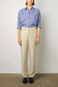 A person stands facing forward, wearing a blue and white vertical striped button-up shirt tucked into a pantalon sable—a light beige pantalon tailleur femme that exudes effortless elegance—paired with black shoes, against a plain background.