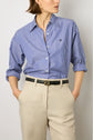 A person wearing a blue and white striped button-up shirt with the sleeves rolled up, paired with a pantalon sable and a black belt, hands in pockets, standing against a plain background.