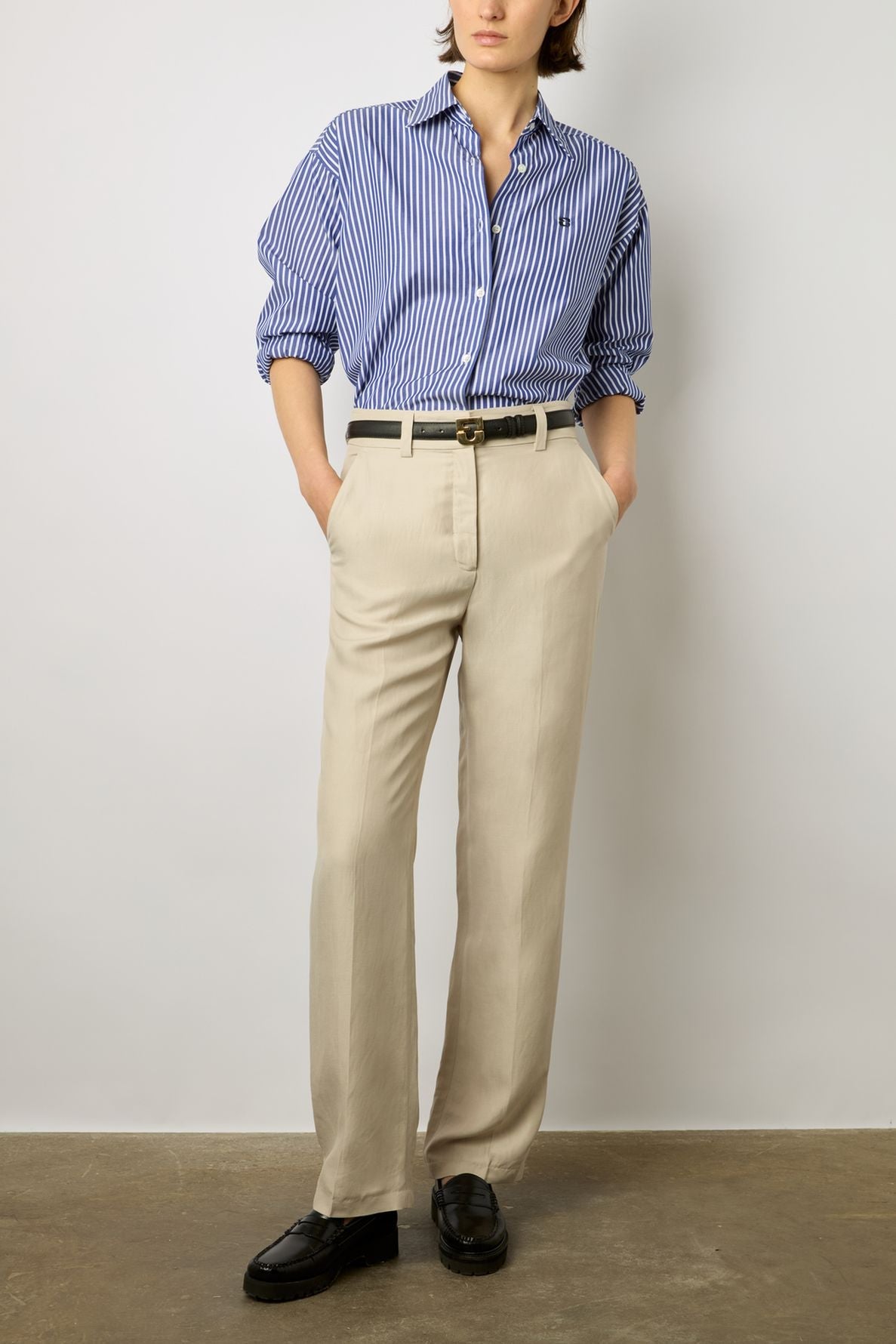 Person wearing a blue and white striped button-up shirt tucked into a pantalon sable with a black belt and black shoes, standing against a plain light background. Hands are in pockets.