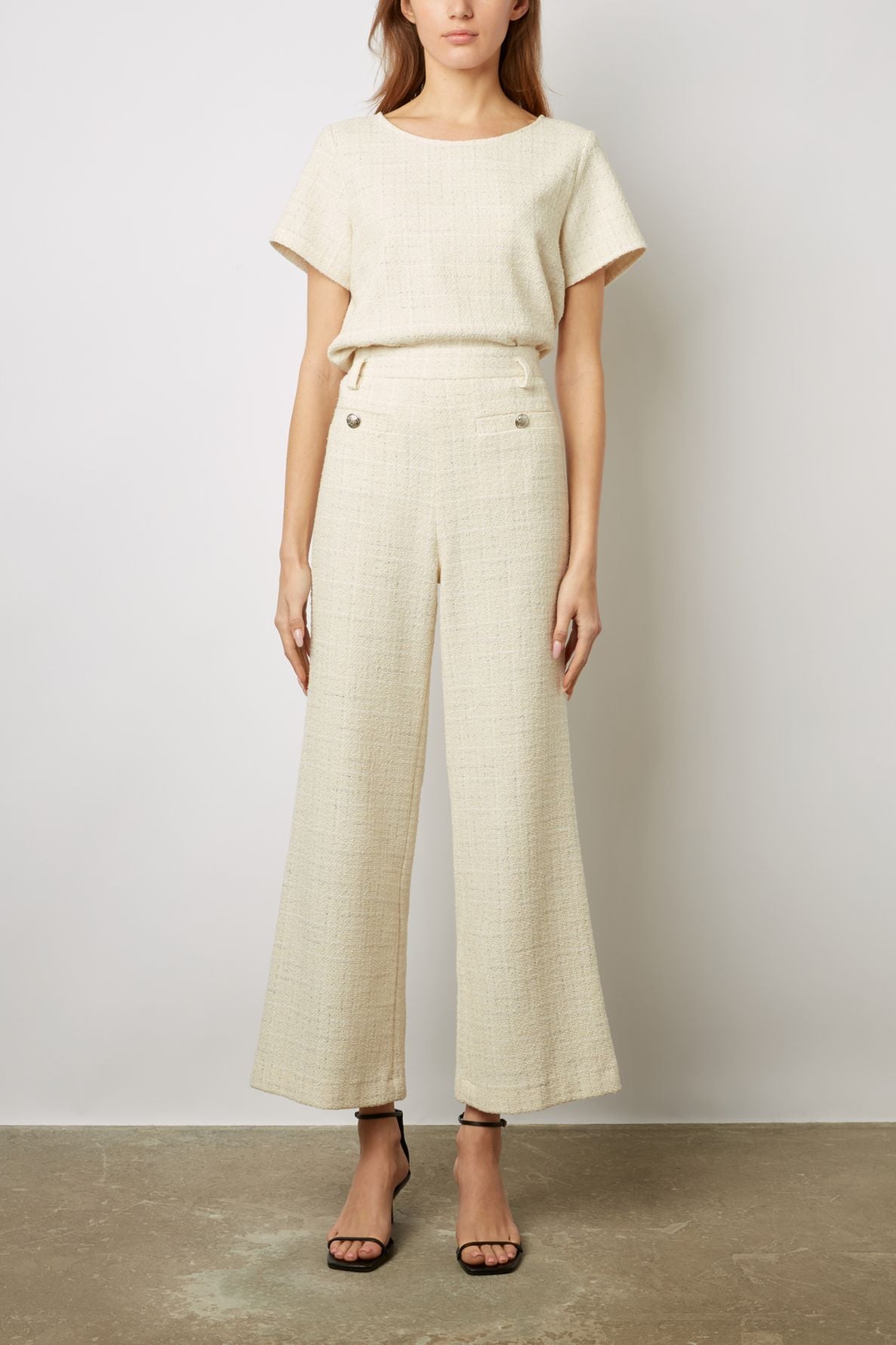 Woman wearing a short-sleeved cream textured top and matching wide-leg pants with button details, paired with black heeled sandals, standing against a plain background.