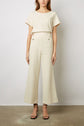 Woman wearing a short-sleeved cream textured top and matching wide-leg pants with button details, paired with black heeled sandals, standing against a plain background.