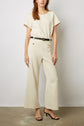 Woman in a cream, short-sleeve top and matching wide-leg pants, black belt, and black high-heeled sandals, standing against a plain background. Her head is partially out of frame.