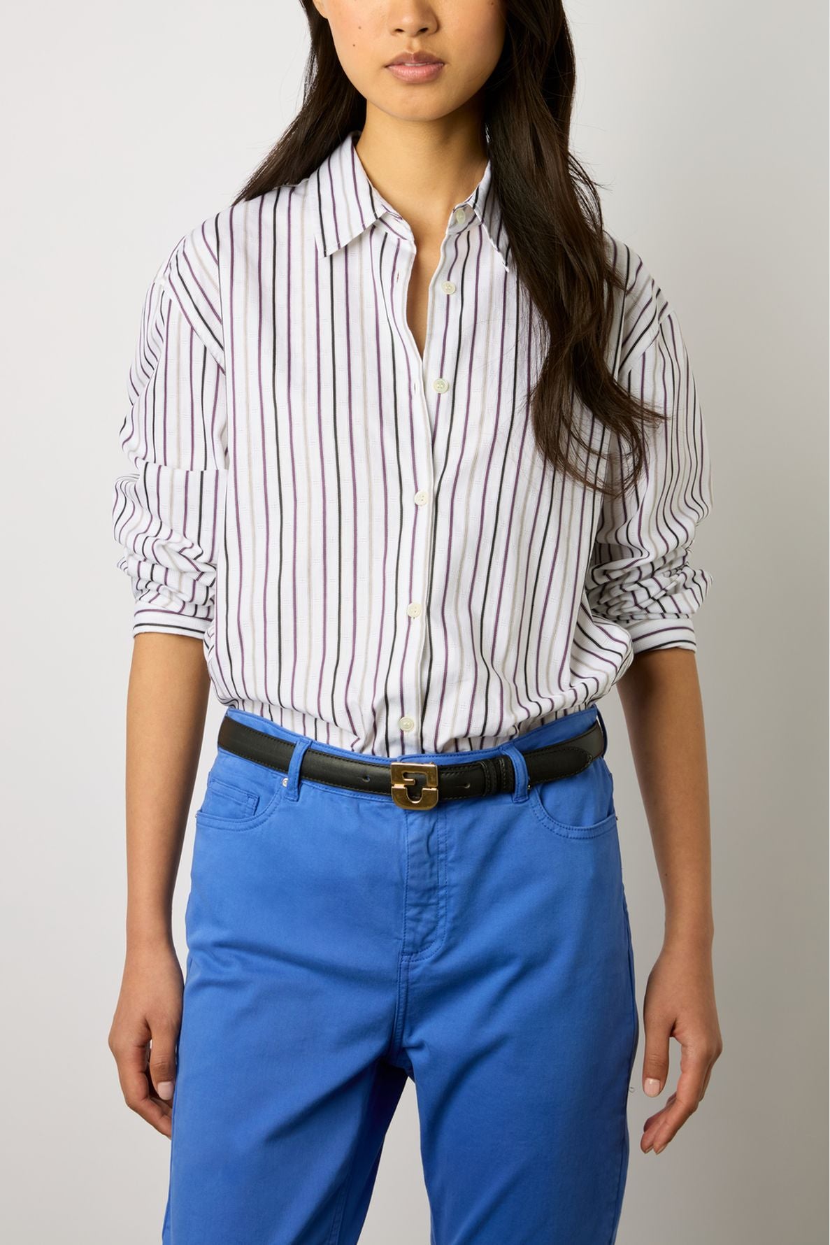 A person wearing a white button-up shirt with vertical maroon stripes, tucked into bright blue pantalon cinq poches with a black belt, standing against a plain background. Their face is partially out of frame.