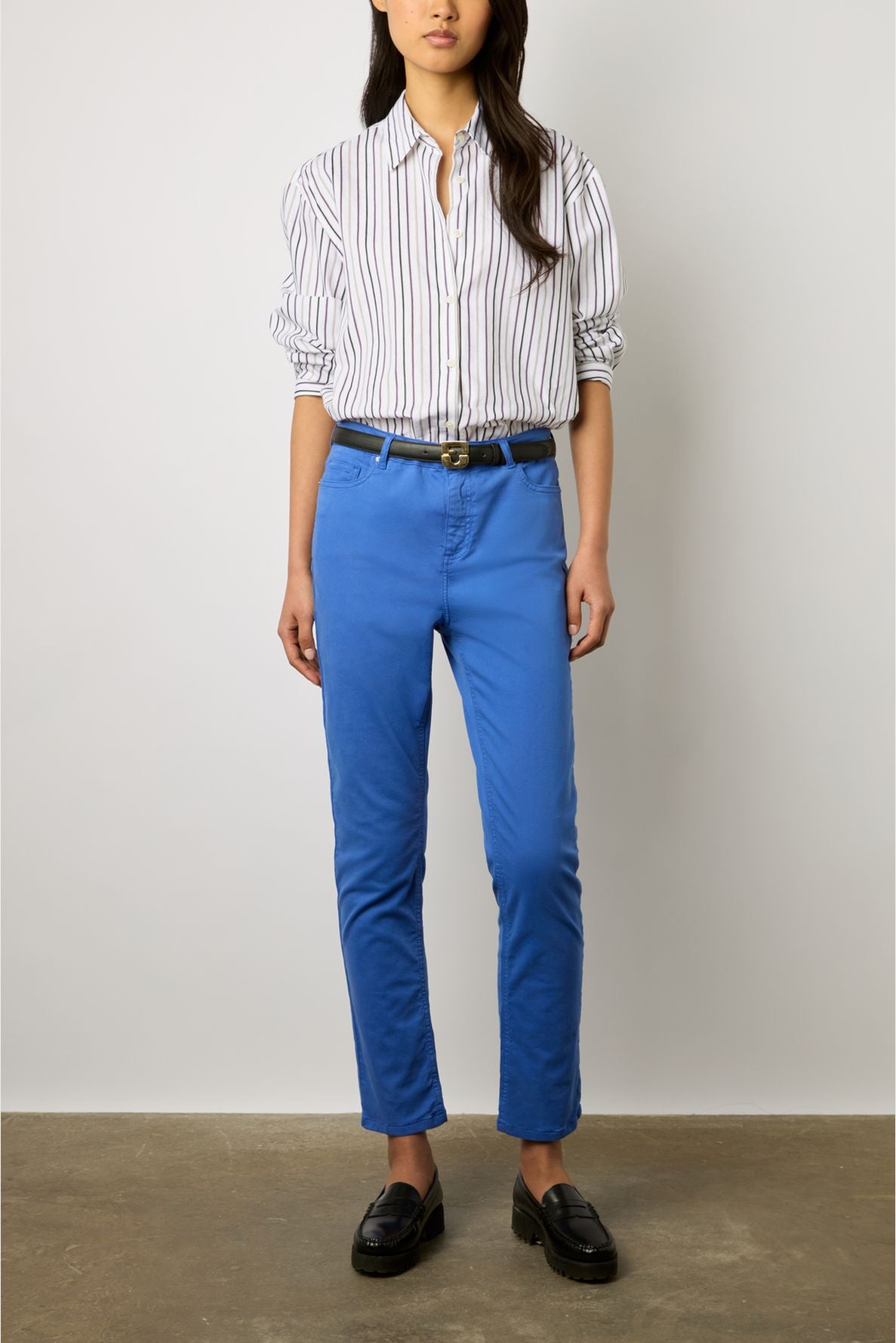 A person stands against a white wall wearing a white shirt with thin black stripes, bright blue denim stretch pants styled in a pantalon slim femme cut, a black belt, and black loafers. The person’s hands are relaxed by their sides.