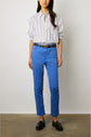 A person stands against a white wall wearing a white shirt with thin black stripes, bright blue denim stretch pants styled in a pantalon slim femme cut, a black belt, and black loafers. The person’s hands are relaxed by their sides.