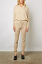 A woman stands against a plain wall wearing a beige hooded jogging cachemire sweater and matching pantalon jogging cachemire with black loafers. Her hands are relaxed by her sides.