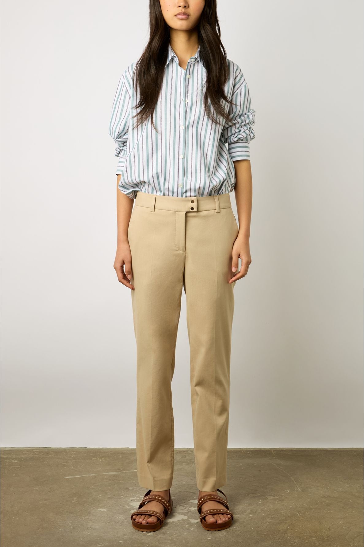 A person stands indoors wearing a striped button-up shirt with rolled sleeves, tan dress pants, and brown studded sandals. Their face is not visible in the image.