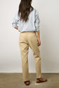 Woman standing with her back to the camera, wearing a striped button-up shirt, beige pants, and brown sandals on a concrete floor against a plain white background.