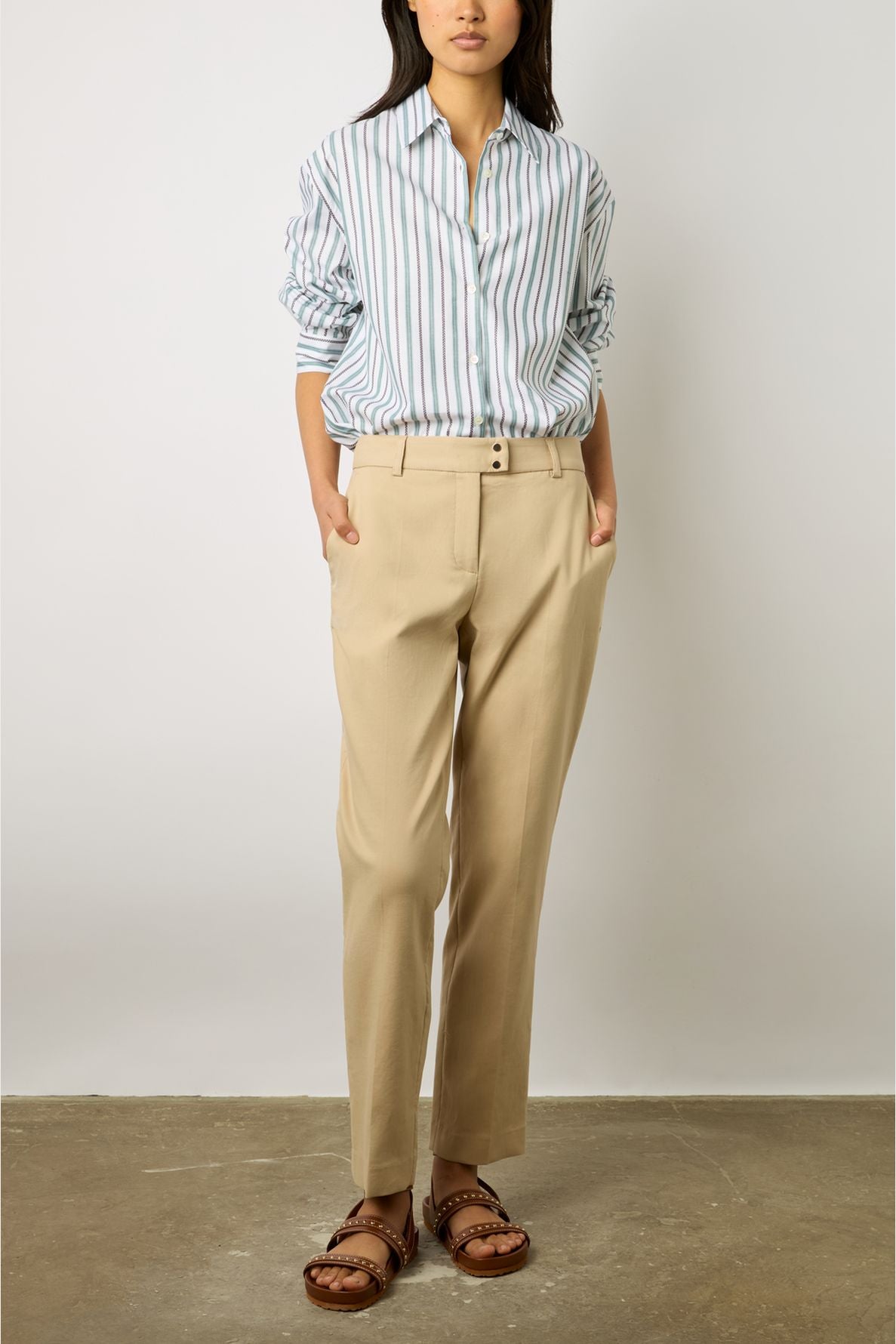 Woman stands against a plain wall wearing a striped button-up shirt, beige pants, and brown sandals with gold studs. Her hands are in her pockets and her sleeves are rolled up. Her face is partially out of frame.