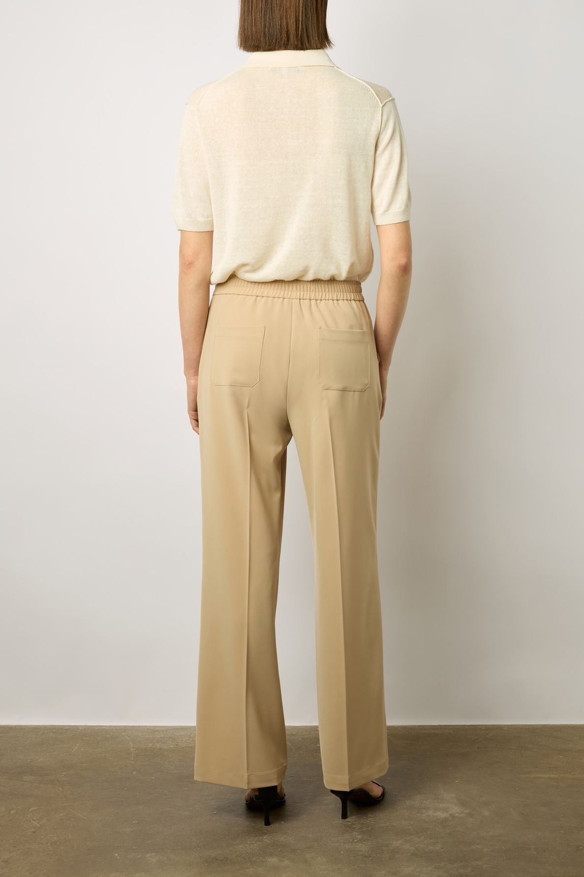 A person stands facing away, wearing a short-sleeved off-white top and high-waisted beige trousers with back pockets. The outfit is paired with black heeled shoes. The background is plain and neutral.