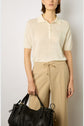 Woman wearing a short-sleeve beige polo shirt and tan drawstring trousers, holding a large black leather handbag by her side, standing against a plain light background.