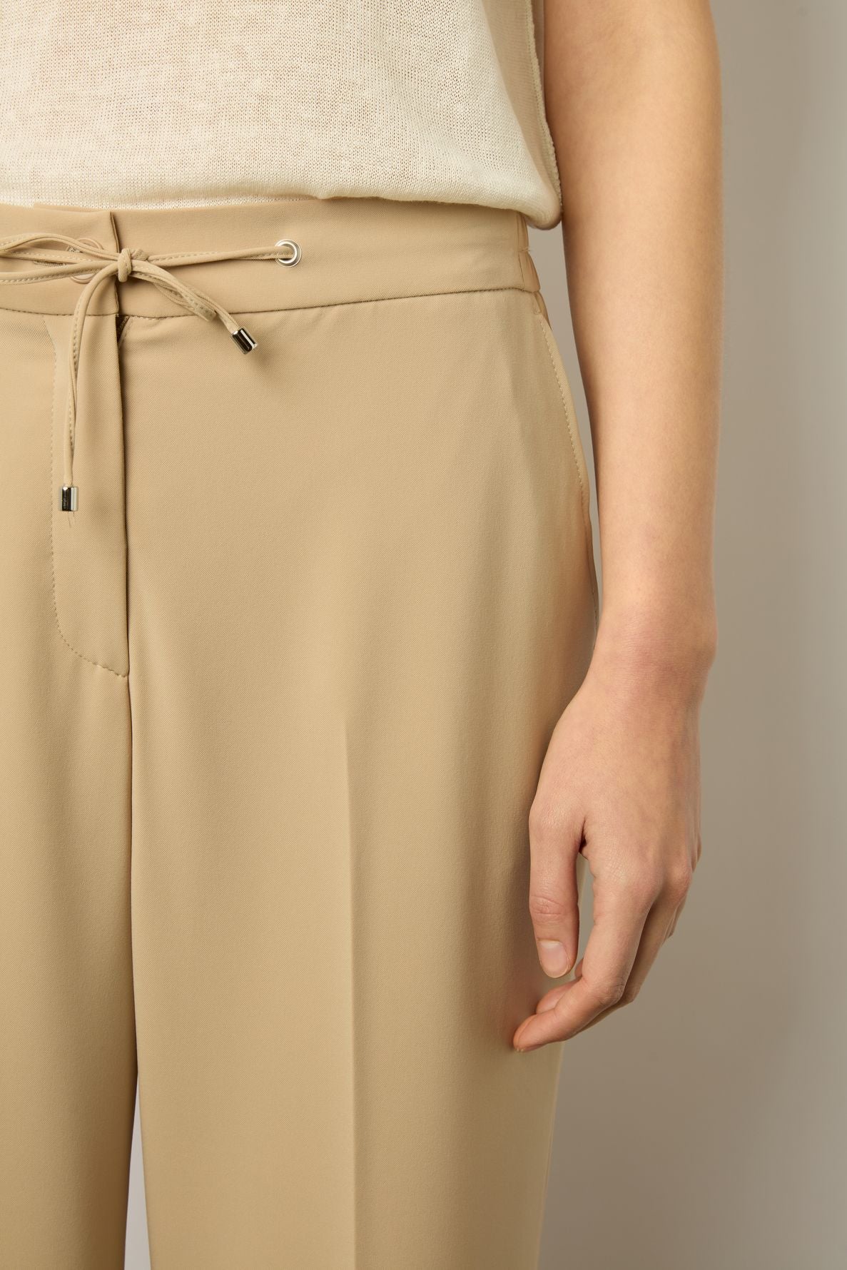 A person wearing beige drawstring trousers and a cream knit top, with their right hand resting alongside their leg. The image focuses on the waist and hip area, with soft lighting.
