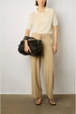 A person wearing a short-sleeved beige polo shirt, tan wide-leg trousers, and black heeled sandals stands against a plain wall, holding a large black handbag.