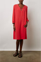A person wearing a knee-length, long-sleeve red robe courte fluide with a col tunisien and black studded sandals stands against a plain background.