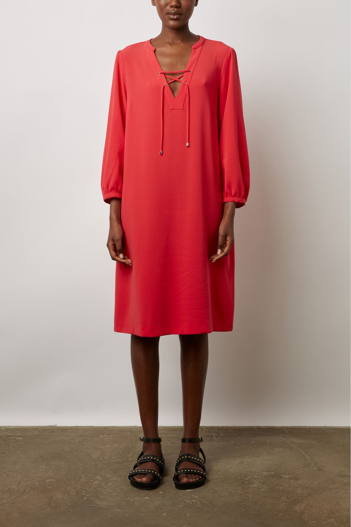 Woman wearing a red, knee-length robe courte fluide with long sleeves and a lace-up neckline style reminiscent of a col tunisien, standing against a plain background. She is also wearing black studded sandals.