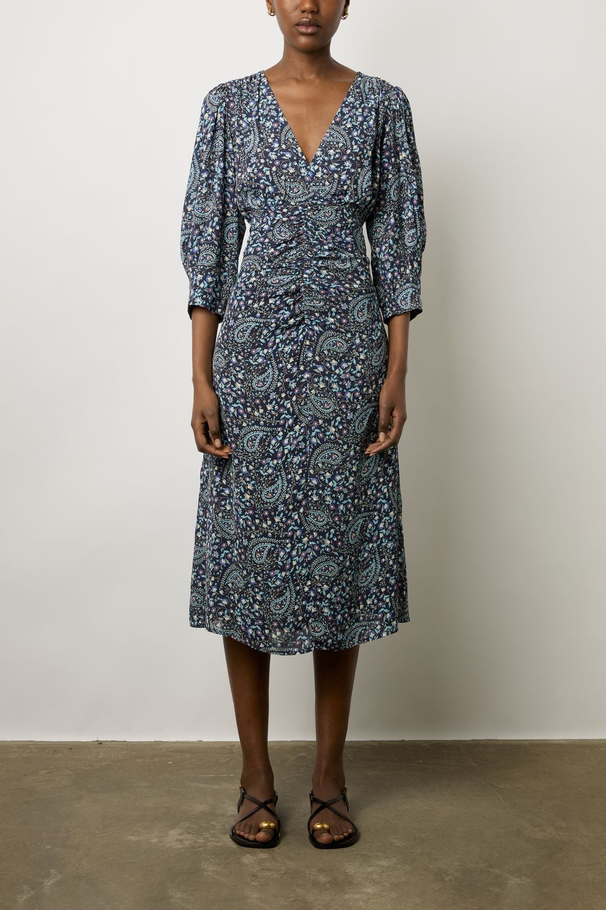 Woman wearing a blue and purple paisley-patterned, knee-length dress with 3/4 sleeves and v-neck, standing against a plain background, wearing brown sandals.