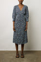 Woman wearing a blue and purple paisley-patterned, knee-length dress with 3/4 sleeves and v-neck, standing against a plain background, wearing brown sandals.