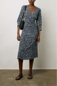 A woman wearing a blue paisley-patterned midi dress with three-quarter sleeves, black flat sandals, and a black shoulder bag stands against a plain wall on a concrete floor.